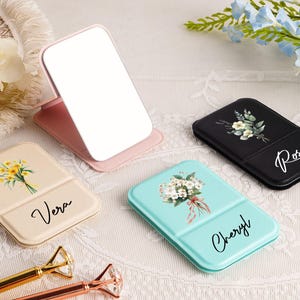 以下が含まれることがあります： ピンク、クリーム、ティールなど、さまざまな色のパーソナライズされたコンパクトミラーのコレクション。各ミラーには、花柄のデザインと「Vera」、「Cheryl」、「Rose」などの名前が特徴です。ミラーは白いレースの表面に表示されています。