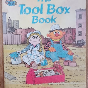 May include: A vintage children's book cover titled "The Tool Box Book" featuring Sesame Street characters. The cover art shows two Muppets, a female and male, with a red toolbox and paint buckets. The book is illustrated by Richard Brown.