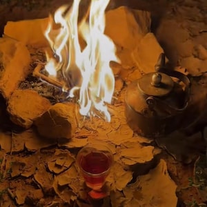 May include: A campfire scene with a bright flame, a vintage metal kettle, and a glass of red liquid. The fire is surrounded by rocks, creating a warm, inviting ambiance. The scene evokes a sense of outdoor relaxation.