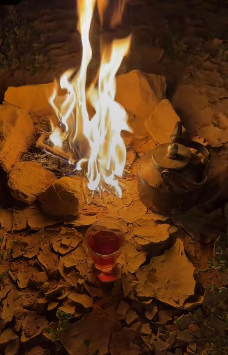 May include: A campfire scene with a bright flame, a vintage metal kettle, and a glass of red liquid. The fire is surrounded by rocks, creating a warm, inviting ambiance. The scene evokes a sense of outdoor relaxation.