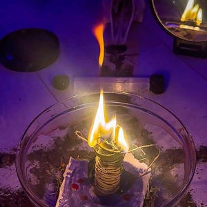 May include: A lit sage bundle in a glass bowl, with a bright flame. The image has a dark, mystical aesthetic, with a goat skull and a mirror in the background. The scene is illuminated by a warm, flickering light.