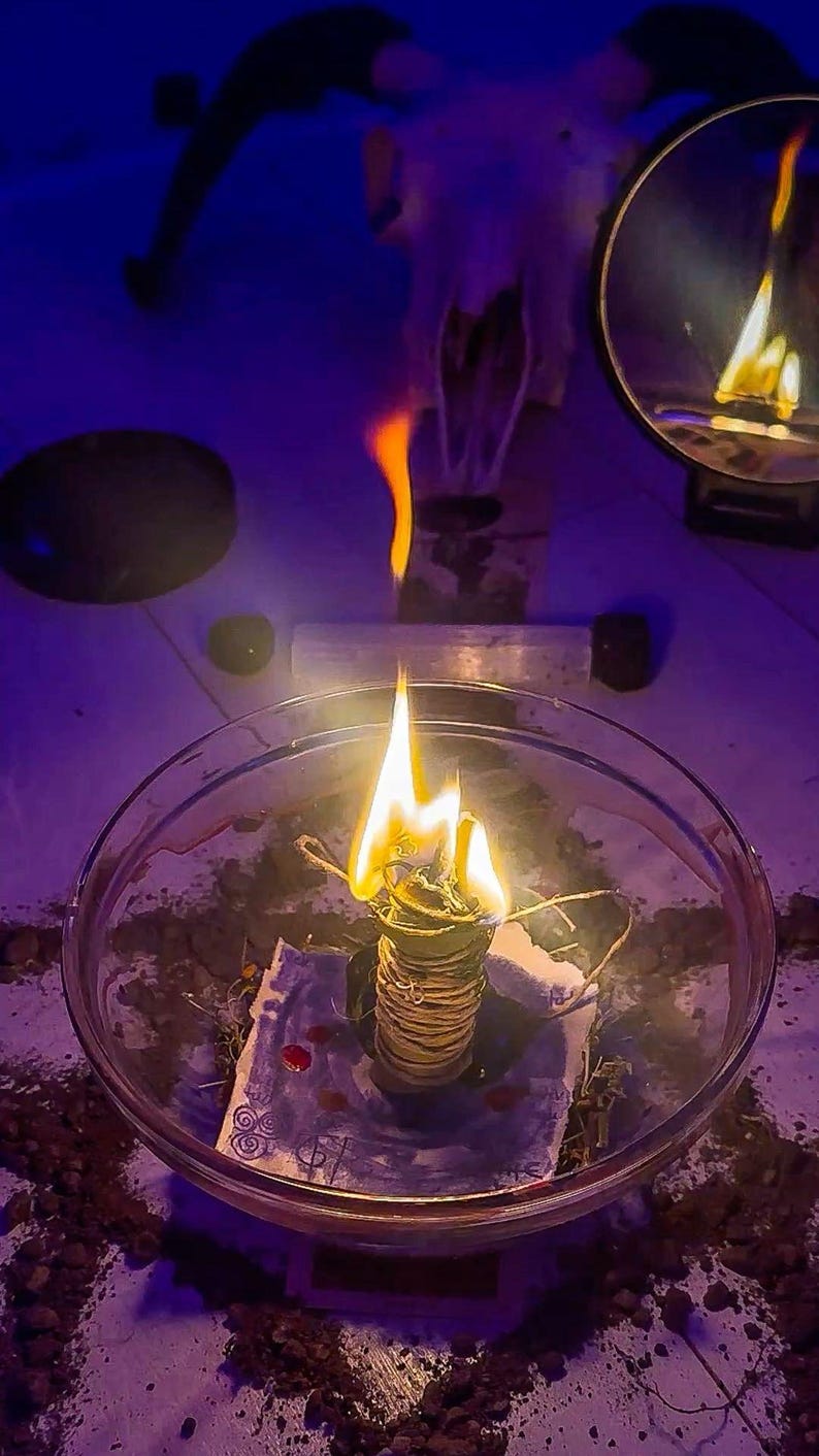May include: A lit sage bundle in a glass bowl, with a bright flame. The image has a dark, mystical aesthetic, with a goat skull and a mirror in the background. The scene is illuminated by a warm, flickering light.