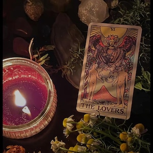 Puede incluir: Una naturaleza muerta oscura con una vela roja encendida, una carta del tarot "THE LOVERS" y flores secas. La carta muestra dos figuras bajo un &aacute;ngel. Cristales y hierbas est&aacute;n dispuestas alrededor de la escena.