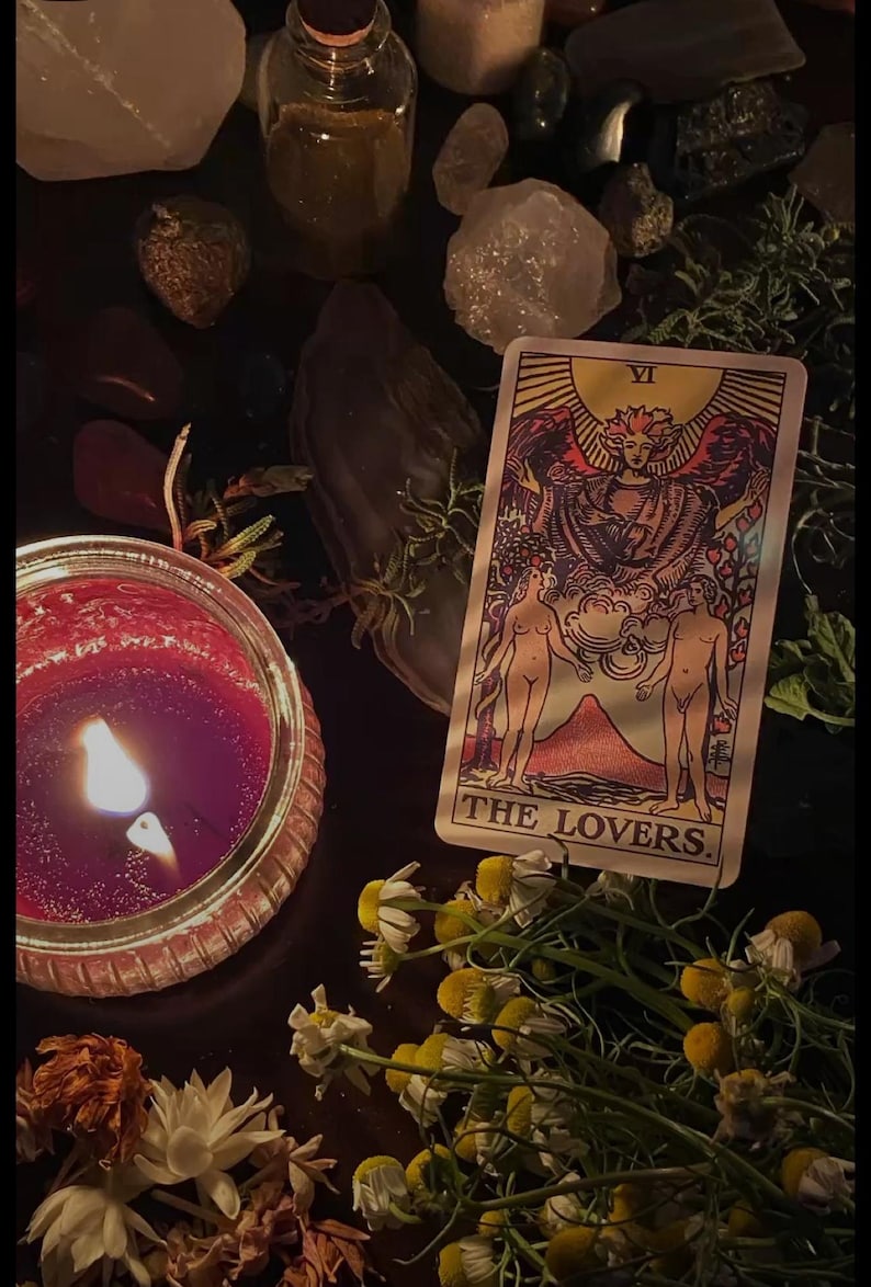 Puede incluir: Una naturaleza muerta oscura con una vela roja encendida, una carta del tarot "THE LOVERS" y flores secas. La carta muestra dos figuras bajo un &aacute;ngel. Cristales y hierbas est&aacute;n dispuestas alrededor de la escena.