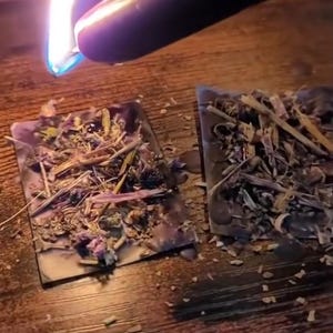 May include: A black candle being lit with a flame, positioned above two rectangular, dried herb-filled squares. The squares are filled with a variety of dried herbs and flowers. The background is a wooden surface.