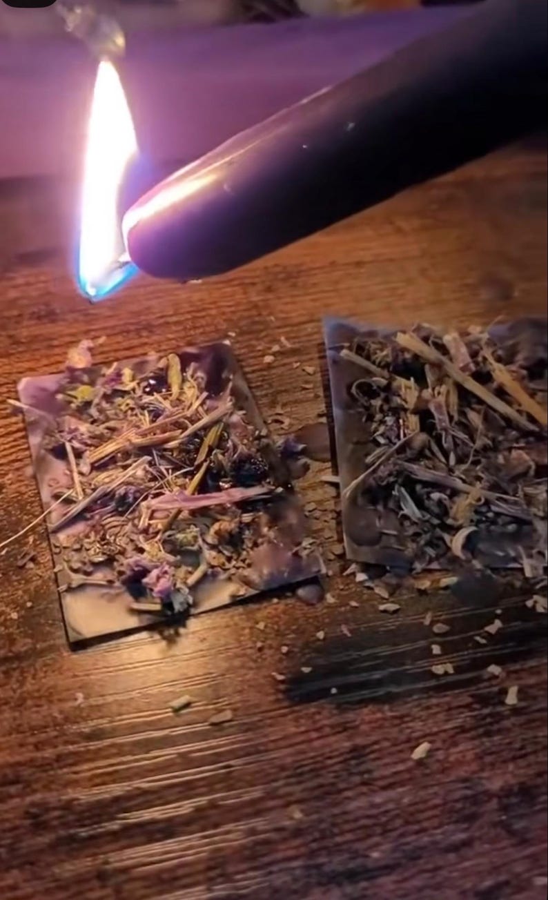 May include: A black candle being lit with a flame, positioned above two rectangular, dried herb-filled squares. The squares are filled with a variety of dried herbs and flowers. The background is a wooden surface.