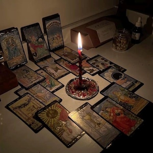 Puede incluir: Una vela con un dise&ntilde;o en espiral rojo y negro arde en medio de cartas del tarot. Las cartas est&aacute;n dispuestas sobre una superficie blanca, con un peque&ntilde;o plato plateado en la base de la vela. Tambi&eacute;n se ven un tarro, una botella y una caja.