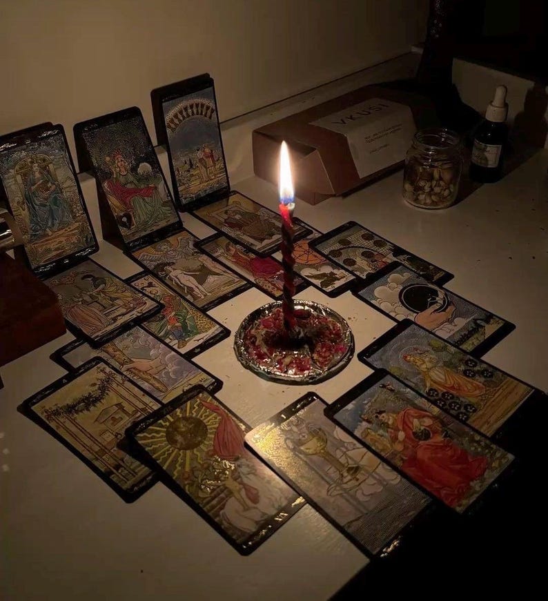 Puede incluir: Una vela con un dise&ntilde;o en espiral rojo y negro arde en medio de cartas del tarot. Las cartas est&aacute;n dispuestas sobre una superficie blanca, con un peque&ntilde;o plato plateado en la base de la vela. Tambi&eacute;n se ven un tarro, una botella y una caja.