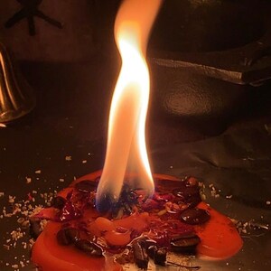 May include: A lit red candle with a tall, bright orange flame. The candle is surrounded by melted wax, herbs, and other small items. The background is dark, with a symbol visible on the left.