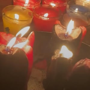 May include: A collection of lit candles in various colors, including red, yellow, and blue, creating a warm, inviting ambiance. The flames flicker, casting shadows and highlighting the wax. The candles are in glass holders.