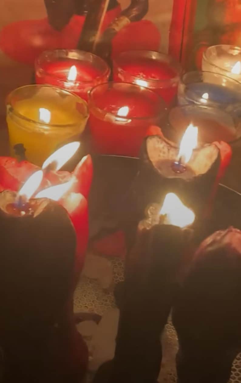 May include: A collection of lit candles in various colors, including red, yellow, and blue, creating a warm, inviting ambiance. The flames flicker, casting shadows and highlighting the wax. The candles are in glass holders.