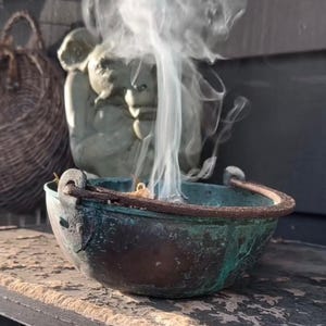 May include: A weathered, copper-toned cauldron with a handle, emitting a stream of white smoke. The cauldron sits on a distressed wooden surface. In the background, a stone statue and a woven basket are visible.