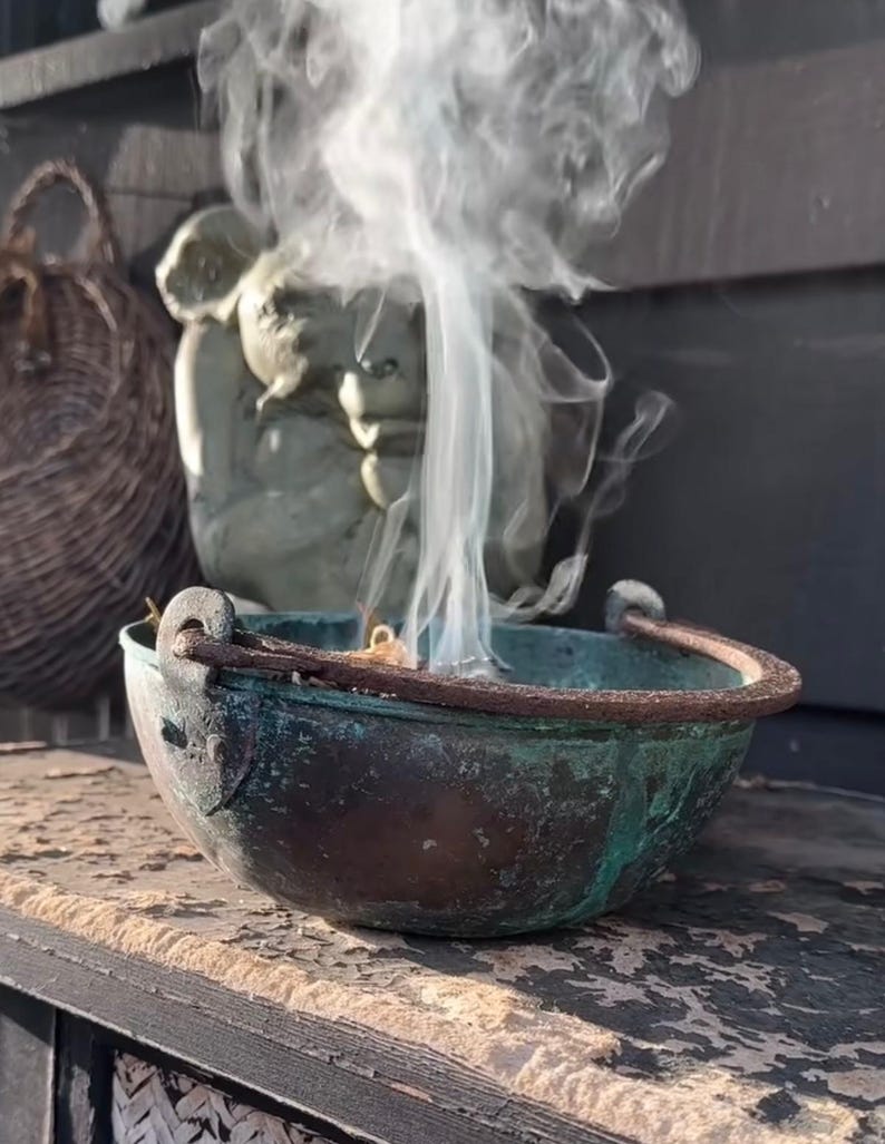 May include: A weathered, copper-toned cauldron with a handle, emitting a stream of white smoke. The cauldron sits on a distressed wooden surface. In the background, a stone statue and a woven basket are visible.