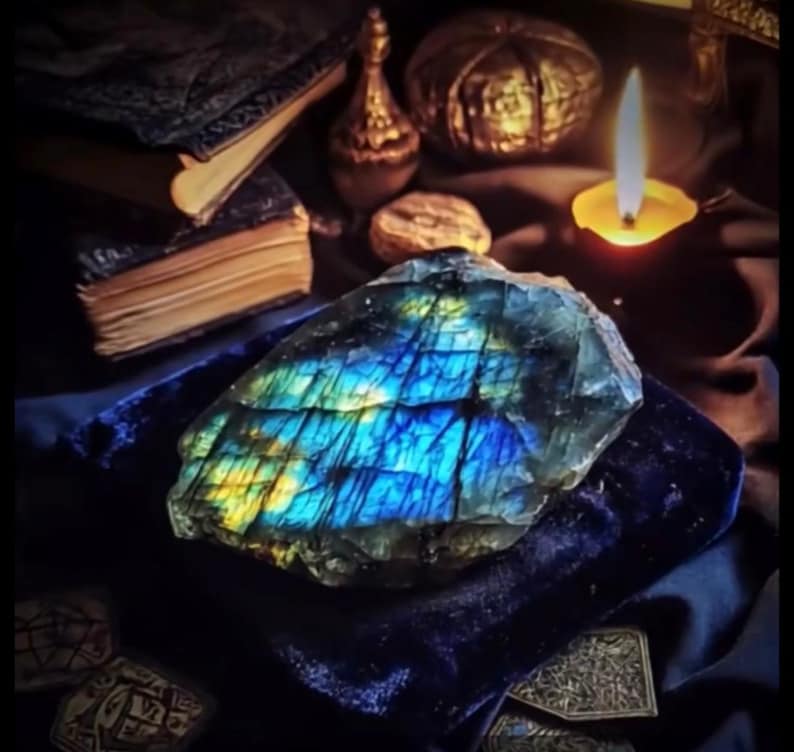 May include: A large, iridescent labradorite gemstone with blue, yellow, and green flashes rests on a dark blue velvet surface. The gemstone is the focal point, surrounded by antique books, a lit candle, and decorative objects, creating a mystical ambiance.