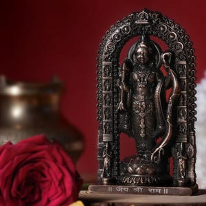 May include: A bronze-coloured statue of a deity, standing within an ornate archway. The statue is on a wooden base with text. A red rose and a brass pot are in the blurred background. The statue is a religious item.