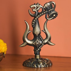 May include: A bronze-coloured decorative statue featuring a trident design, an Om symbol, and a hammer-like object. The statue has intricate detailing and stands on a textured base. The background is a warm orange colour.