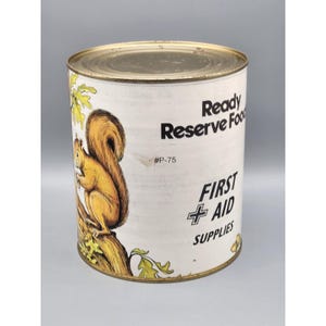 May include: Vintage metal can with a gold lid and base, featuring a cartoon squirrel illustration. The can is labeled "Ready Reserve Food" and "First Aid Supplies" in black text. The squirrel is brown and orange, perched on a tree branch.