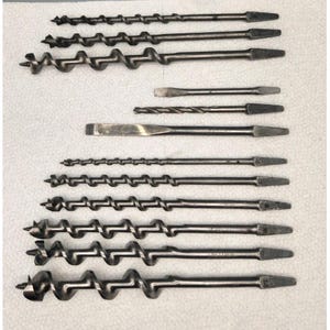 May include: A collection of various metal drill bits and a screwdriver are arranged on a white surface. The drill bits vary in size and style, with spiral and straight designs. The tools are silver and black.