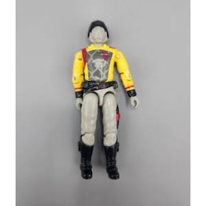 May include: A vintage action figure of a soldier in a yellow uniform with black boots and a black helmet. The figure has gray legs and arms, and a red and gray emblem on the chest. The figure is standing upright.