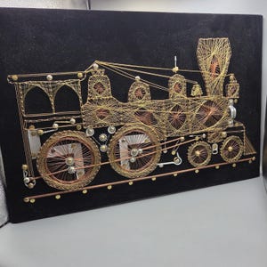May include: A string art wall piece on a black background depicting a steam locomotive. The train is constructed from gold and copper-colored wire wrapped around metal nails, featuring intricate geometric patterns, circular wheels, and a tall smokestack.