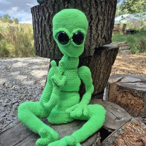 May include: A handmade, green crocheted alien doll with large black eyes. The alien is seated with its legs crossed and arms outstretched. The doll is made of soft yarn and is sitting on a wooden surface.