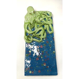 May include: A ceramic wall hanging featuring a green octopus with yellow eyes and black spots on its tentacles. The octopus is set against a blue background with colorful speckles, creating a marine theme.