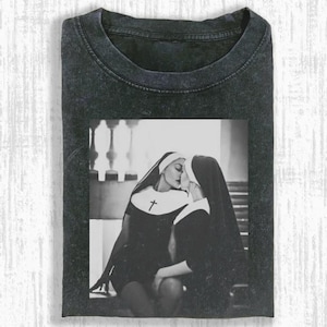 May include: Dark gray t-shirt featuring a black and white image of two nuns kissing. The image is centered on the shirt and depicts the nuns in traditional habits. The t-shirt has a crew neck and short sleeves.