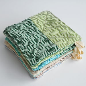 May include: A stack of green, blue, and beige kitchen towels with a diamond pattern. The towels are folded and have a fringe edge.