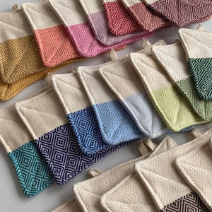May include: A collection of colorful, diamond-patterned pot holders. Each pot holder features a natural-colored base with a contrasting band of color, including yellow, orange, pink, red, blue, green, and purple. The pot holders have a loop for hanging.