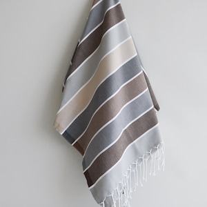 Puede incluir: Una toalla a rayas en tonos marrones, grises y beige con flecos blancos. La toalla est&aacute; colgada en una pared blanca.