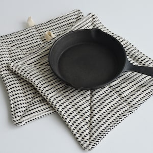 May include: A black cast iron skillet resting on two square quilted pot holders. The pot holders feature a white fabric base with a repeating pattern of small, diagonal black dashes and include small beige fabric loops for hanging.