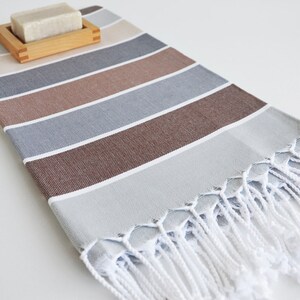 Puede incluir: Una toalla de mano a rayas con un esquema de color marr&oacute;n, gris y beige. La toalla tiene flecos blancos y est&aacute; hecha de algod&oacute;n. Una pastilla de jab&oacute;n est&aacute; sentada en un jabonera de madera.