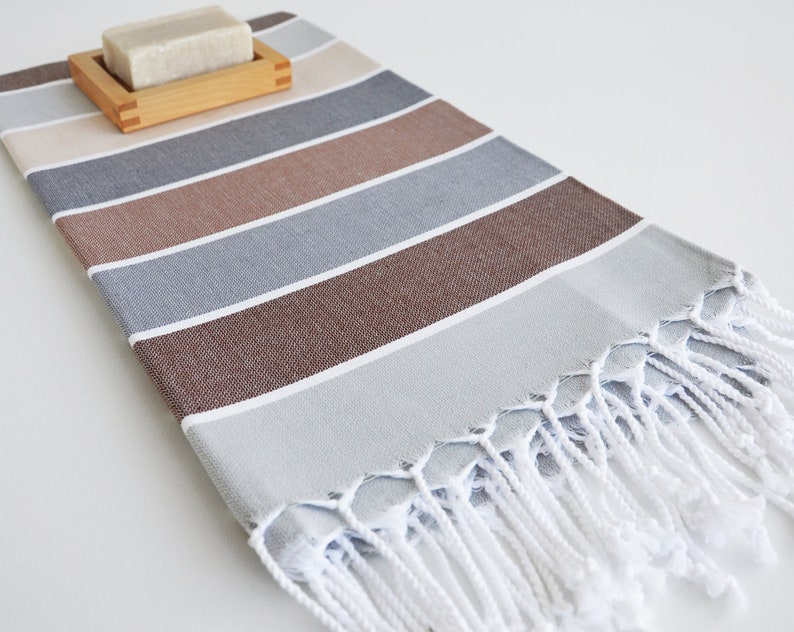 Puede incluir: Una toalla de mano a rayas con un esquema de color marr&oacute;n, gris y beige. La toalla tiene flecos blancos y est&aacute; hecha de algod&oacute;n. Una pastilla de jab&oacute;n est&aacute; sentada en un jabonera de madera.