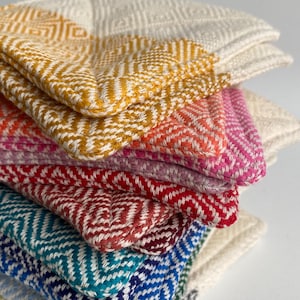 May include: A stack of woven fabric coasters featuring a geometric diamond pattern. The coasters are layered in various colors including mustard yellow, orange, pink, red, teal, and blue, all contrasted against a white woven base thread.