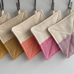 May include: Five square pot holders with a cream-colored top and a patterned bottom in shades of gold, brown, orange, pink, and purple. Each pot holder is hanging from a metal hook.