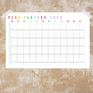May include: A white 2026 year planner with a colorful header and monthly columns. The text "YEAR PLANNER 2026" is at the top, with months labeled across the top. The planner is taped to a textured, tan wall.