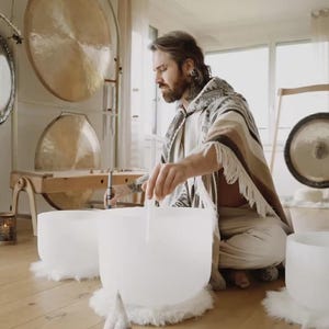 Puede incluir: Una persona está sentada en un suelo de madera, tocando cuencos cantores de cristal. La habitación tiene varios gongs de diferentes tamaños. La persona lleva un chal estampado y pantalones blancos. Los cuencos son blancos y están sobre alfombras blancas y esponjosas.