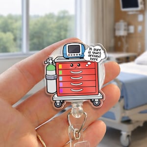 May include: A badge reel featuring a cartoon medical cart with a monitor, oxygen tank, and the text "It sure is quiet around here." The cart is red with drawers and sits on wheels. The reel has a clear plastic clip and a retractable cord.