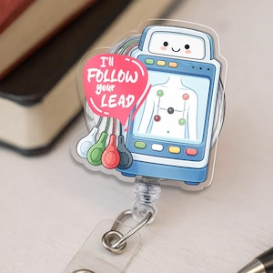 May include: A blue and white medical-themed badge reel featuring a cartoon EKG machine with a smiling face. A pink speech bubble reads "I'll Follow Your Lead." The reel has a clear plastic strap and a metal clip.