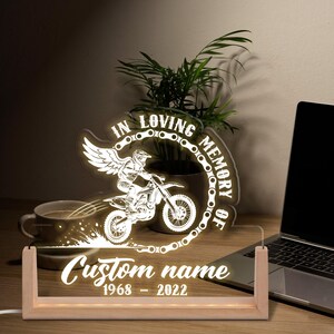 May include: A lighted memorial sign with a motocross rider design. The sign reads "In Loving Memory Of" and includes a space for a custom name and dates. The sign is made of clear acrylic and wood.