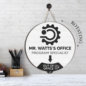May include: A round, wooden office sign with a white face and black accents. The sign reads "MR. WATTS'S OFFICE PROGRAM SPECIALIST" and has a rotating "OUT OF OFFICE" indicator. The sign hangs from a rope.