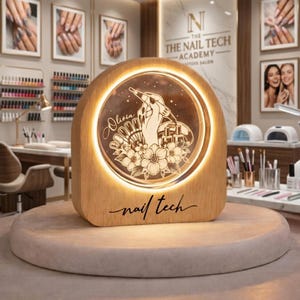 May include: A wooden, illuminated nail tech sign with a circular design featuring a hand holding a nail tool, flowers, and the name "Olivia." The sign is on a round, light-colored pedestal, with the words "nail tech" at the bottom.