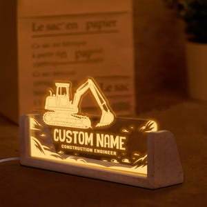 May include: A lighted desk nameplate with a wooden base. The nameplate features a glowing excavator design and the text "CUSTOM NAME CONSTRUCTION ENGINEER". The light is a warm yellow color.