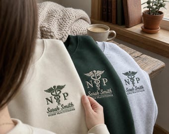 May include: Three crewneck sweatshirts in cream, dark green, and heather gray. Each features an embroidered caduceus symbol above the letters NP, the name Sarah Smith, and the text Nurse Practitioner in a dark green or black thread.