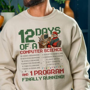 May include: Beige sweatshirt with a green and red pixelated design. The text reads "12 Days of a Computer Science" with a Santa Claus graphic at a computer. The list includes debugging, servers humming, and emails sending.