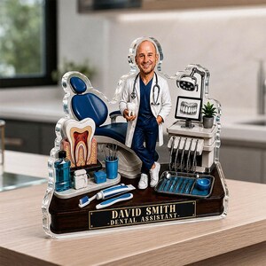 May include: A custom acrylic figurine of a dental assistant in a white coat and blue scrubs, standing next to a dental chair, a tooth model, dental tools, and a monitor. The base features a plaque with the text DAVID SMITH - DENTAL ASSISTANT.