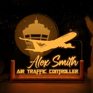 May include: A glowing orange acrylic desk lamp featuring an airplane and air traffic control tower design. The lamp displays the name "Alex Smith" and the title "AIR TRAFFIC CONTROLLER" in a stylized font. The lamp has a wooden base.