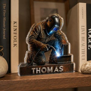 May include: A decorative acrylic display featuring a kneeling welder wearing a protective helmet and brown work gear. The figure is actively welding a metal piece, emitting a bright blue spark. The base is dark brown with the name THOMAS in silver.