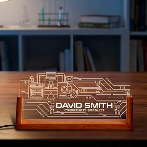 May include: A lighted desk nameplate with a circuit board design. The nameplate is made of clear acrylic and sits in a wooden base. The name "DAVID SMITH" and title "CYBERSECURITY SPECIALIST" are displayed in white.