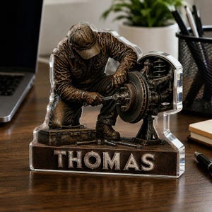 May include: A bronze-toned figurine of a mechanic kneeling while working on a car brake assembly with a wrench. The figure is set on a rectangular base featuring the name THOMAS in raised silver lettering, all encased in a clear acrylic frame.